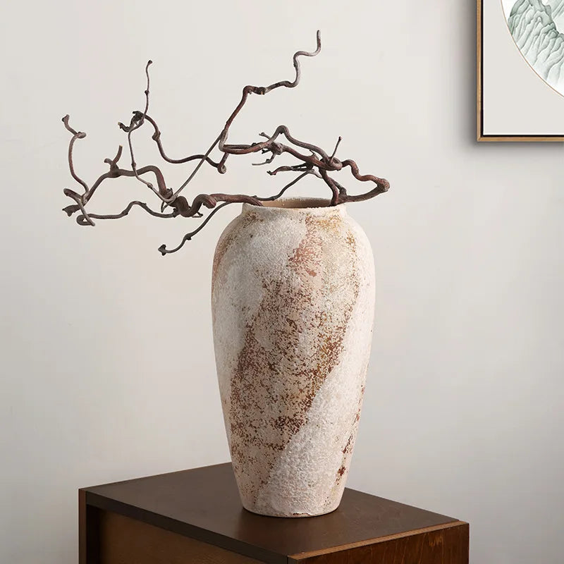 Tall weathered clay jar with a sculptural branch on a wooden stool