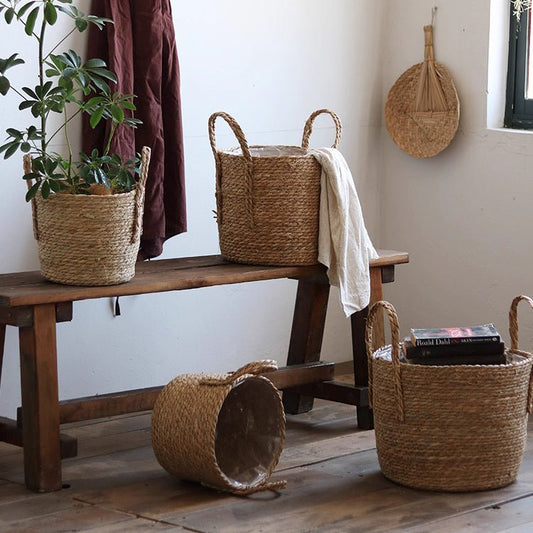 Three handwoven seagrass baskets with rope handles, styled with a plant and linen on a wood bench